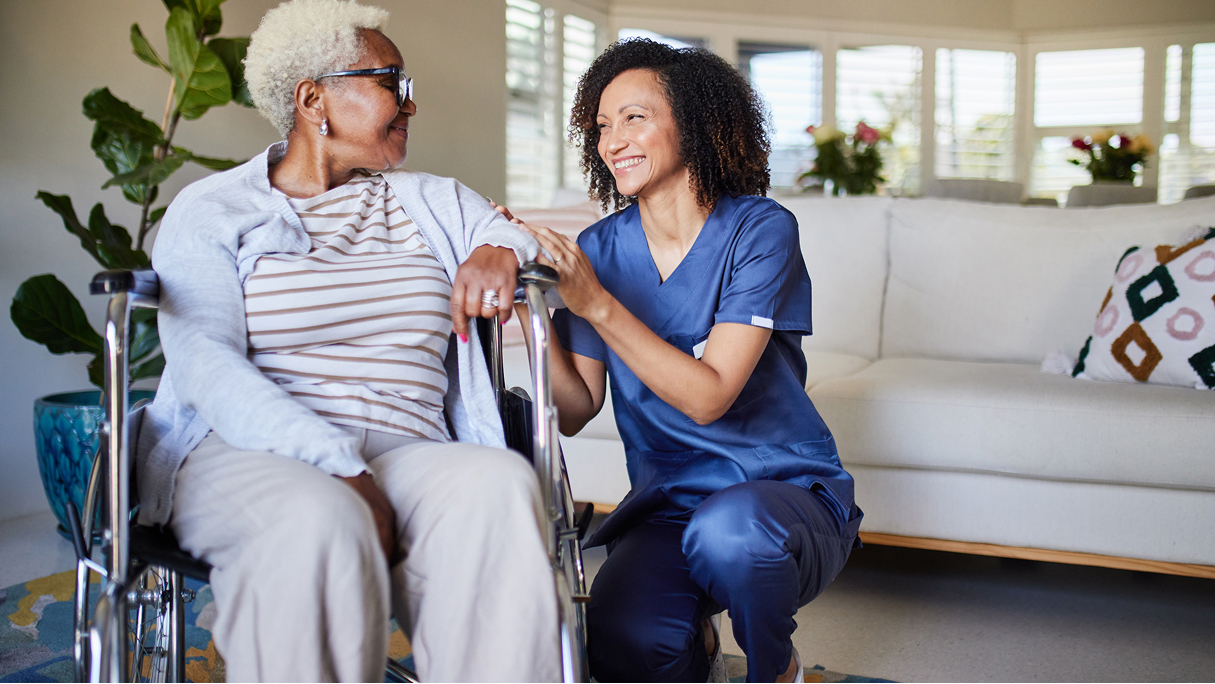 A healthcare worker smiles and chats with an older woman in a wheelchair in a living room, showing medbridge patient care.