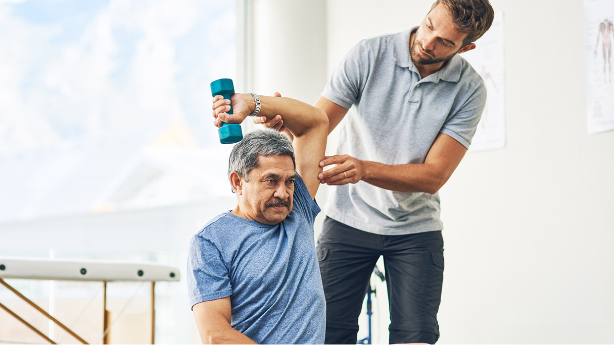 A man does a dumbbell arm exercise with a physical therapist in a clinic using digital healthcare tools like medbridge.