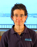 Individual with short curly hair and glasses, in a dark polo shirt, standing indoors with a window view of water.