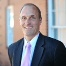 Smiling man in a suit and pink tie stands before a brick wall.