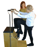 An elderly woman uses a wooden step machine for exercise with a trainer's assistance in medbridge training.