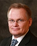 A confident man in glasses, a suit, and a patterned tie represents the future of digital healthcare before a dark textured background.