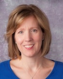 A smiling woman with light brown hair and a blue top is against a gray background.