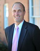 A man in a suit and pink tie smiles outside a brick building, representing digital healthcare professionalism for Medbridge.