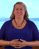 A blonde person in a blue top and necklace is shown with a blurred background.