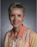 A person with short gray hair smiles in a light blouse with an orange collar, set against a gray professional background, possibly Medbridge.