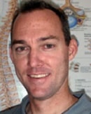 A smiling person with short hair stands in front of a medical diagram, showcasing their Medbridge healthcare expertise.