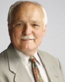 An older man in a light gray suit and patterned tie smiles slightly, against a plain background.