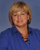 A person with short blond hair, wearing a blue collared shirt and earrings, smiles against a plain blue background, embodying the seamless integration of medbridge into daily life.