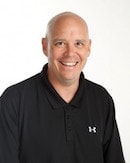 A smiling person with a shaved head in a black Under Armour polo against a white background represents digital healthcare innovation at medbridge.