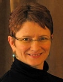 A smiling person with short brown hair, glasses, and a black top is set against a warm brown background, representing an interest in digital healthcare and medbridge.