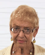 An older person with short, light hair and glasses wears a beige jacket against a plain background.