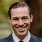 Person in a suit smiles outdoors with a blurred natural background.