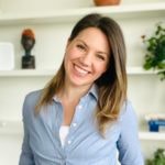 A smiling woman in a blue shirt stands before shelves with plants and decor, embodying medbridge's fresh digital healthcare approach.