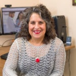A woman with curly hair and a gray sweater sits smiling at a desk with digital healthcare tools, with medbridge technology visible in the background.