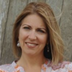 A person with blonde hair in a patterned blouse smiles in front of a wooden backdrop, symbolizing hybrid care.