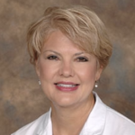 A smiling individual with short blond hair in a white shirt, possibly participating in medbridge healthcare training against a blurred brown backdrop.
