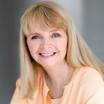 A person with long blonde hair smiles in an orange top against a neutral background.