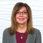 A smiling woman with glasses, brown hair, and a maroon top stands against a white textured wall.