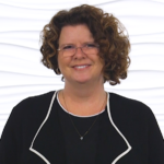 A smiling individual with curly hair and glasses, dressed professionally, stands against a white background, symbolizing medbridge's healthcare training.