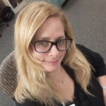 A woman with long blonde hair and glasses, wearing a dark top, sits thoughtfully, contemplating innovative digital healthcare solutions with a slight smile.