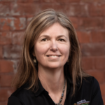 A long-haired individual in a black shirt smiles in front of a red brick wall, representing medbridge's digital healthcare spirit.