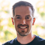 A bearded person with short hair smiles against a blurred green background, embodying digital healthcare harmony in medbridge.