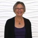 A smiling individual with glasses, wearing a necklace and black cardigan over a purple top, stands against a white background, embodying medbridge's blend of digital healthcare and warmth.