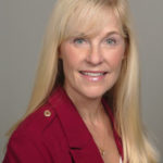 A smiling woman with long blonde hair wearing a red blazer is pictured against a gray background.