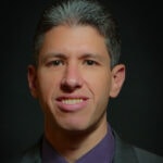 A man with short gray hair in a dark suit smiles warmly, representing modern healthcare advancements like medbridge.