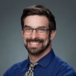 A smiling person with glasses, a beard, and short hair wears a blue shirt and patterned tie against a gray background.