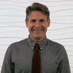 A smiling person with short hair, wearing a gray shirt and red tie, stands confidently, embodying modern healthcare training expertise for medbridge.