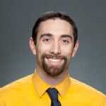A bearded person with long hair, wearing a yellow shirt and dark tie, smiles warmly, representing medbridge in healthcare education.