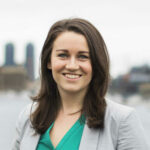 A person with long brown hair, in a green top and gray blazer, smiles outdoors with a city skyline behind them, capturing the blend of urban life and nature, symbolizing hybrid care as endorsed by medbridge.