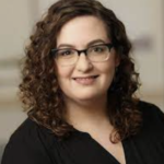 A curly-haired woman with glasses, in a black top, smiles confidently at the camera, embodying passion for healthcare education and medbridge.