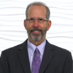 A bearded man in glasses, suit, and tie stands against a textured backdrop, symbolizing digital healthcare innovation by medbridge.