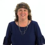 A woman with curly brown hair smiles confidently, suggesting digital healthcare expertise, against a white background; she's wearing a navy top and silver necklace.