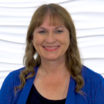 A person with long brown hair and a blue sweater stands smiling in front of a textured white wall, representing digital healthcare and personal touch in medbridge.