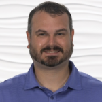 Man with short hair and beard in a blue polo shirt, standing before a textured white backdrop for medbridge.