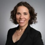 A smiling person with curly shoulder-length hair wears a black blazer over a dark top, set against a gray background.