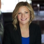 Person smiling with short blonde hair in a black blazer and blue shirt indoors.