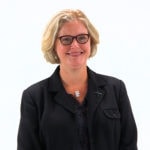 A smiling individual with short blonde hair, glasses, and a black suit stands against a white background, exemplifying Medbridge's professional healthcare training.