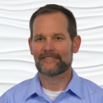 A bearded person in a light blue shirt stands against a wavy white background.