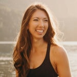 A person with long brown hair smiles by the water in a black tank top.