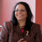A woman with long dark hair and glasses, wearing a brown blazer with an emblem and a pink top, smiles in a red and beige room.