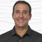 Man with short dark hair in a dark shirt stands against a wavy white background.