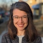 A smiling woman with long brown hair and glasses discusses digital healthcare innovation outdoors in a gray jacket.