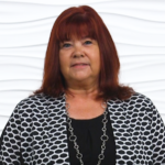 Red-haired woman in a black top and patterned jacket stands professionally against a white textured background, reflecting medbridge's healthcare education environment.