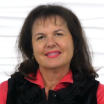A smiling woman with dark hair in a black vest and red shirt stands against a white textured background.