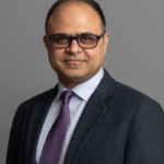 A man with glasses in a suit and tie stands before a gray background at medbridge.